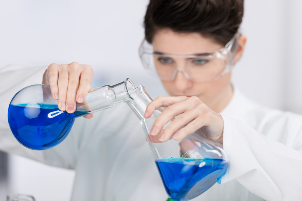 Young woman working in a chemistry laboratory wearing safety goggles and pouring a bright blue chemical solution between conical glass flasks